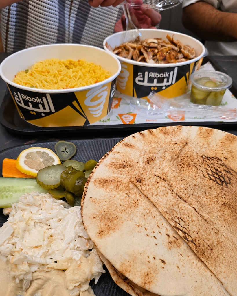 Warmes Fladenbrot im Vordergrund, dahinter Albaik-Schalen mit Chicken Shawarma und gelbem Reis sowie eingelegte Gurken auf dem Tisch bei Shawarma Albaik Berlin